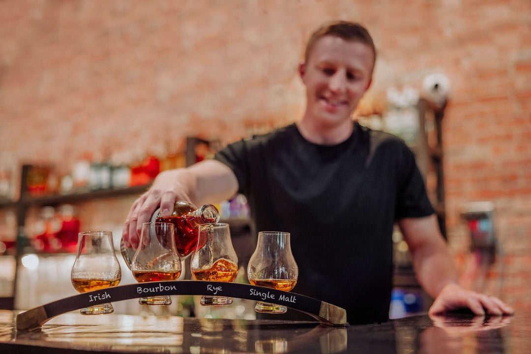 Person holding a tray with glass samples of different types of whiskey in a bar setting.