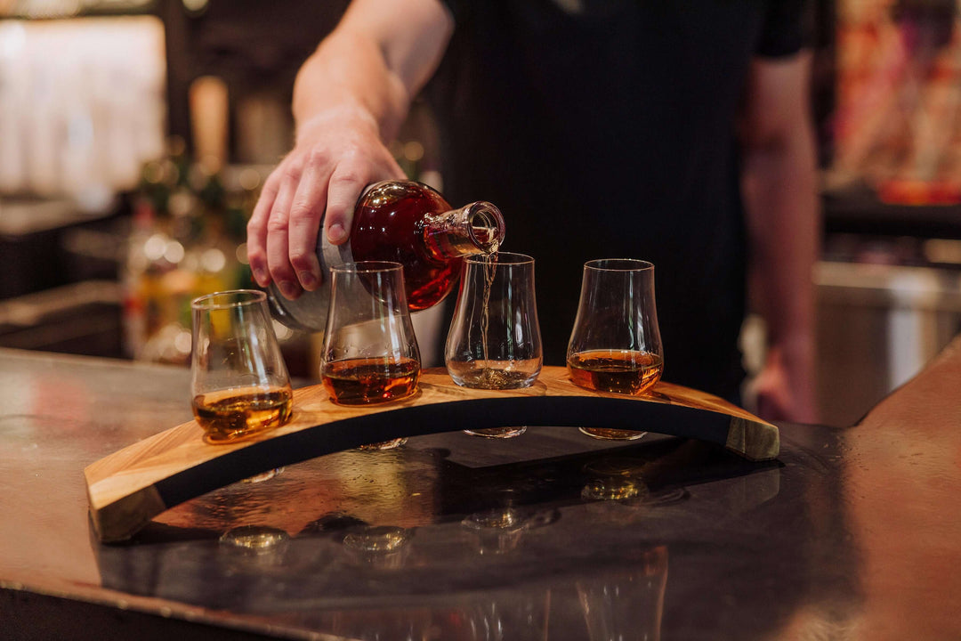 Person pouring whiskey into four glasses on a wooden tray.