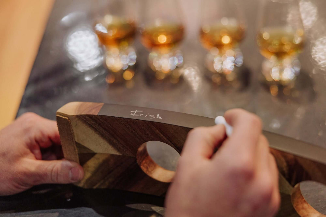 Person holding a wooden board with 'Irish' written on it, in front of blurred glasses of whiskey.