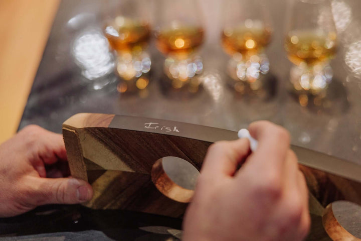 Person holding a wooden board with 'Irish' written on it, in front of blurred glasses of whiskey.