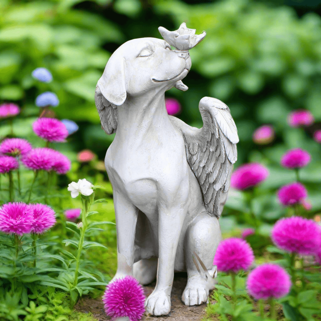 Angelic dog memorial statue with wings, gazing at a bird in vibrant flower garden.