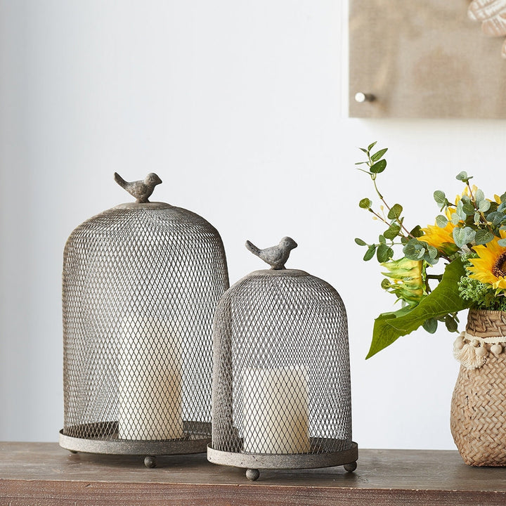 Two wire mesh candle holders with birds on top, next to a vase of flowers on a wooden surface.