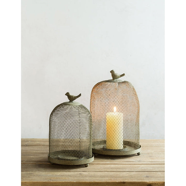 Two decorative mesh candle holders with birds on top on a wooden surface.