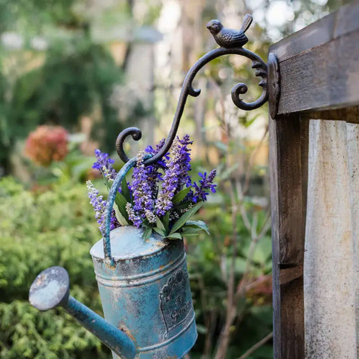 Antique cast iron bird wall hook holding a vintage watering can with purple flowers in a garden setting.