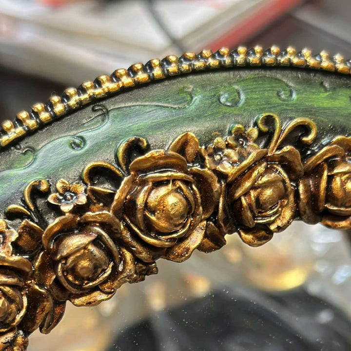 Close-up of ornate gold detailing on an antique green oval wall mirror, featuring intricate rose motifs.