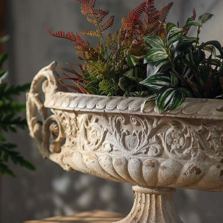 Decorative stone planter with ornate designs filled with artificial plants.