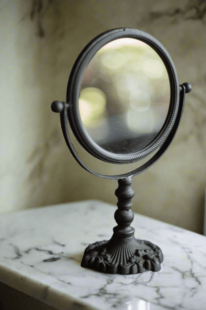 Antique swivel tabletop makeup mirror in ornate cast iron, placed on a marble surface, showcasing vintage elegance.