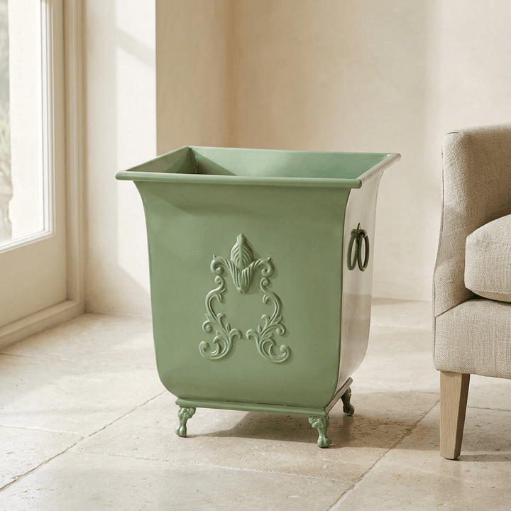 Aveline metal planter in Soft Garden Green with raised scrollwork and decorative feet on a limestone floor beside a linen chair.