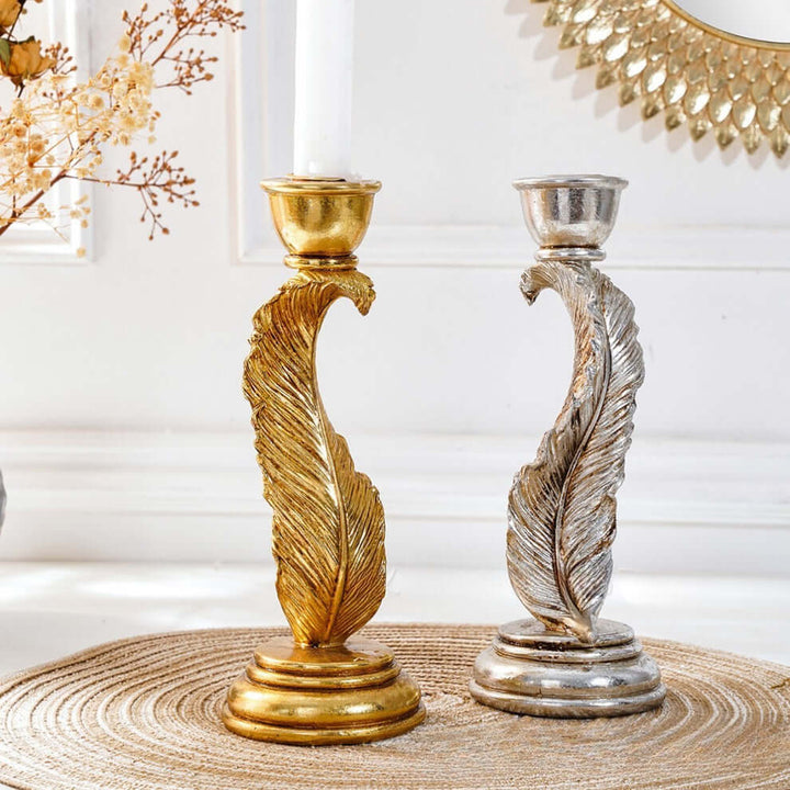 Gold and silver feather-shaped candle holders on a textured surface with a decorative mirror in the background.