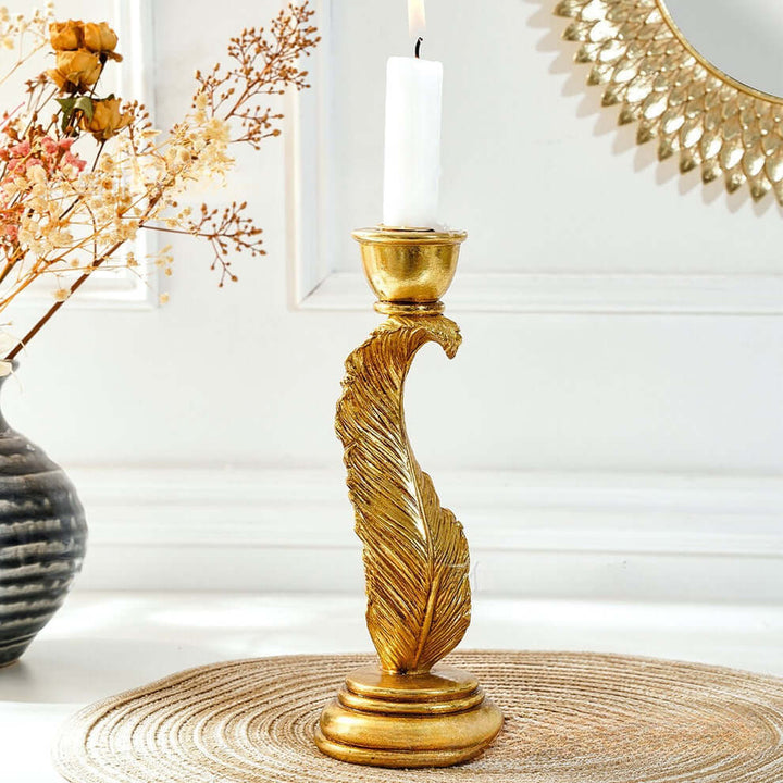 Gold candle holder with a white candle on a table next to a vase with flowers and a decorative mirror.