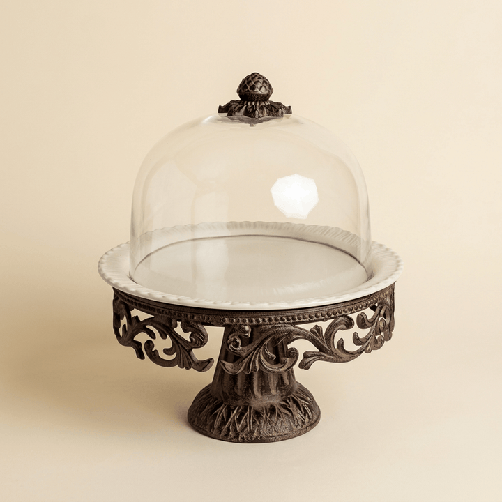 Bellavita cream ceramic cake pedestal with dark acanthus metalwork and glass dome on an ivory background.