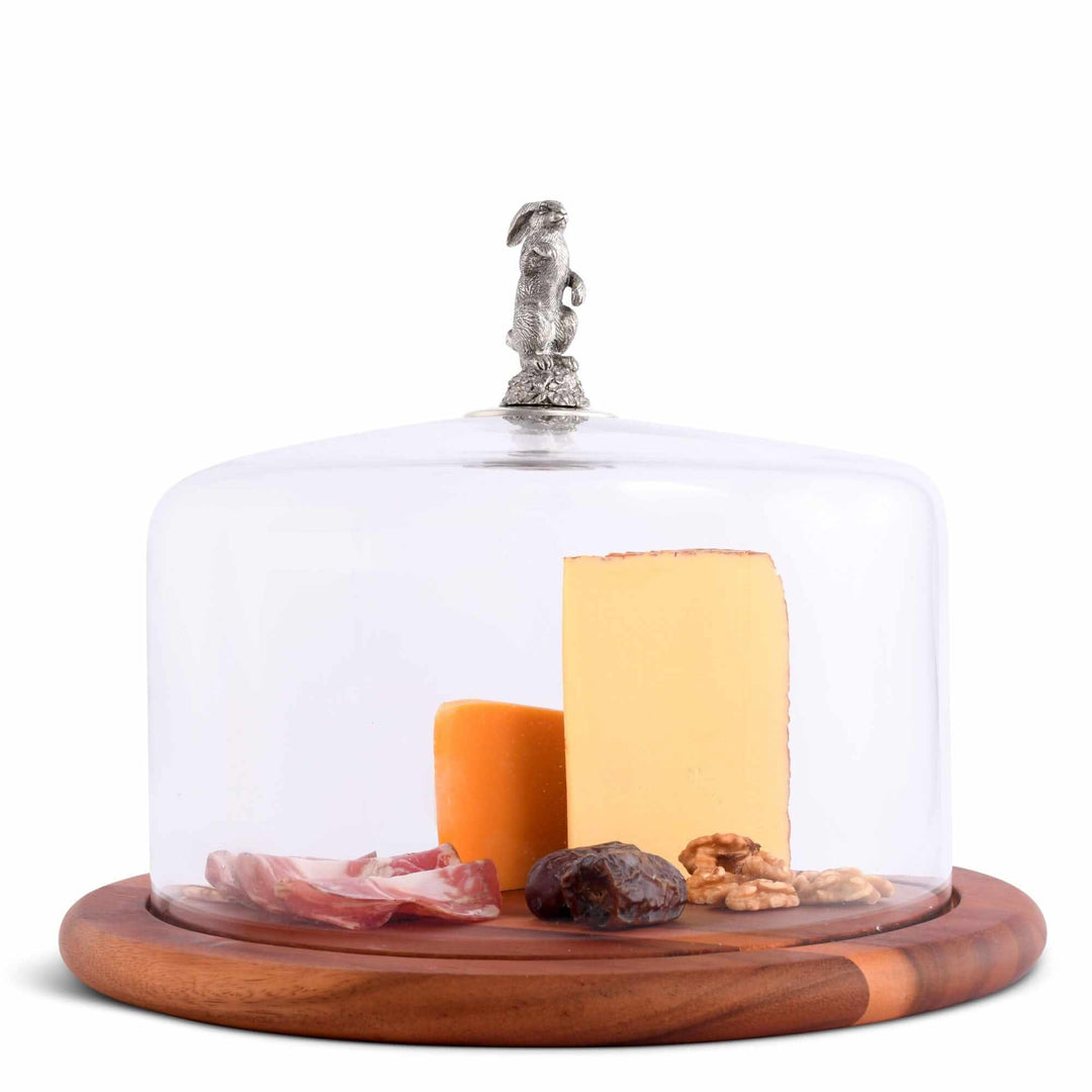 Cheese platter with a glass dome, wooden base, and various cheeses and meats on a white background