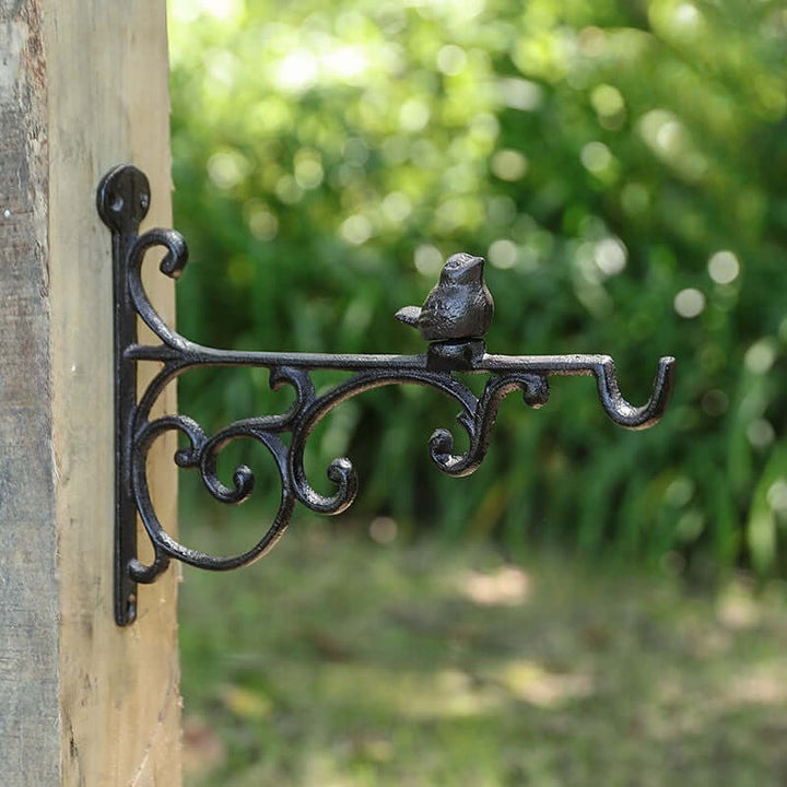 Decorative metal hook with bird design attached to a wooden post, blurred greenery in the background