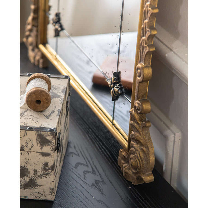 Close-up of vintage arched wall mirror with antique gold frame and floral detailing beside a patterned box on a dark table.