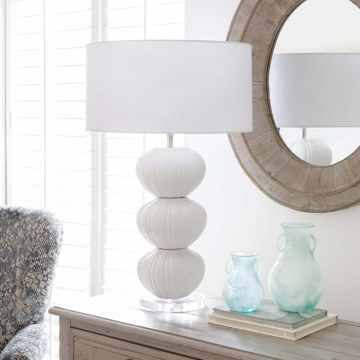 Decorative coastal table lamp with stacked sea urchin base on a wooden surface next to a mirror and vases.
