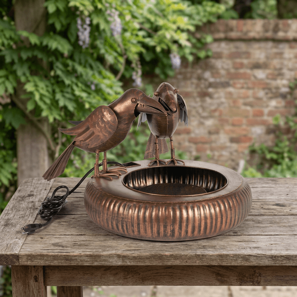 Antiqued Copper Chattering Crows Fountain staged unplugged on a rustic wooden table with greenery and a brick wall behind.