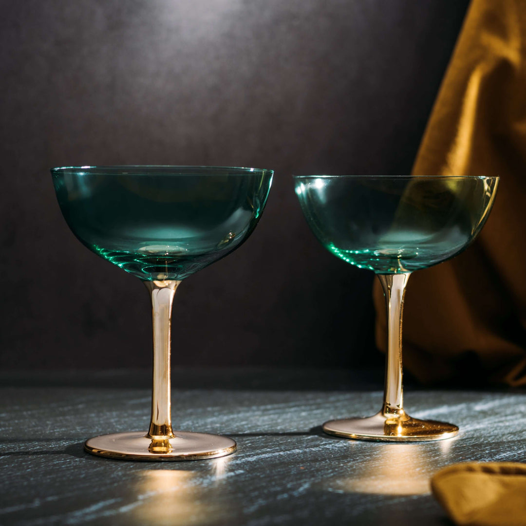 Two green cocktail glasses with gold stems on a dark surface.