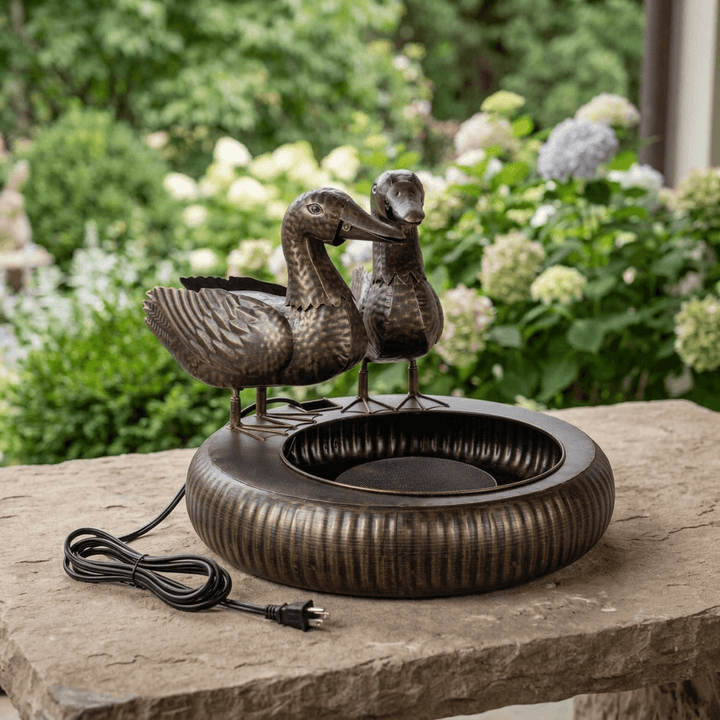 Black Bronze Dabbling Ducks Fountain staged unplugged on a stone table with soft garden greenery and hydrangeas behind.