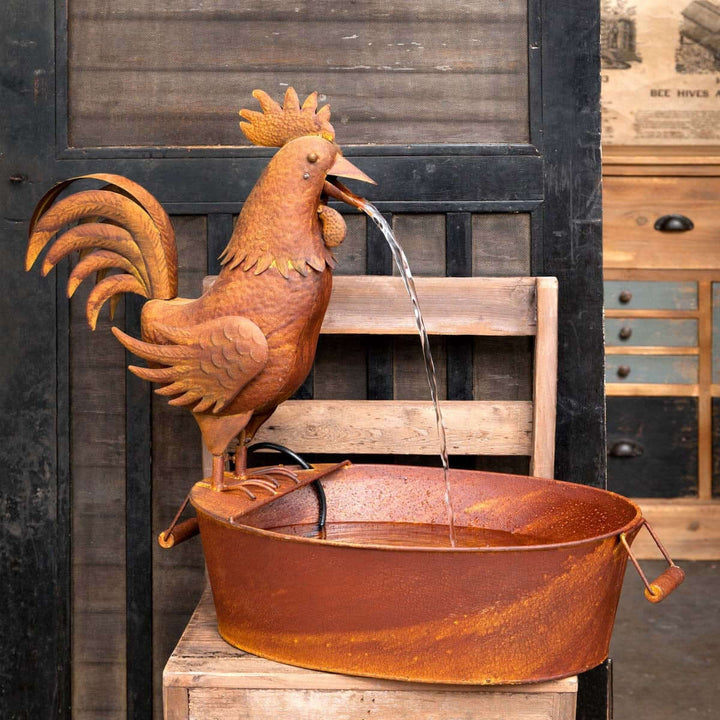 Vintage farmhouse rooster fountain made of rusted metal on a wooden chair, with water trickling into a rustic basin.