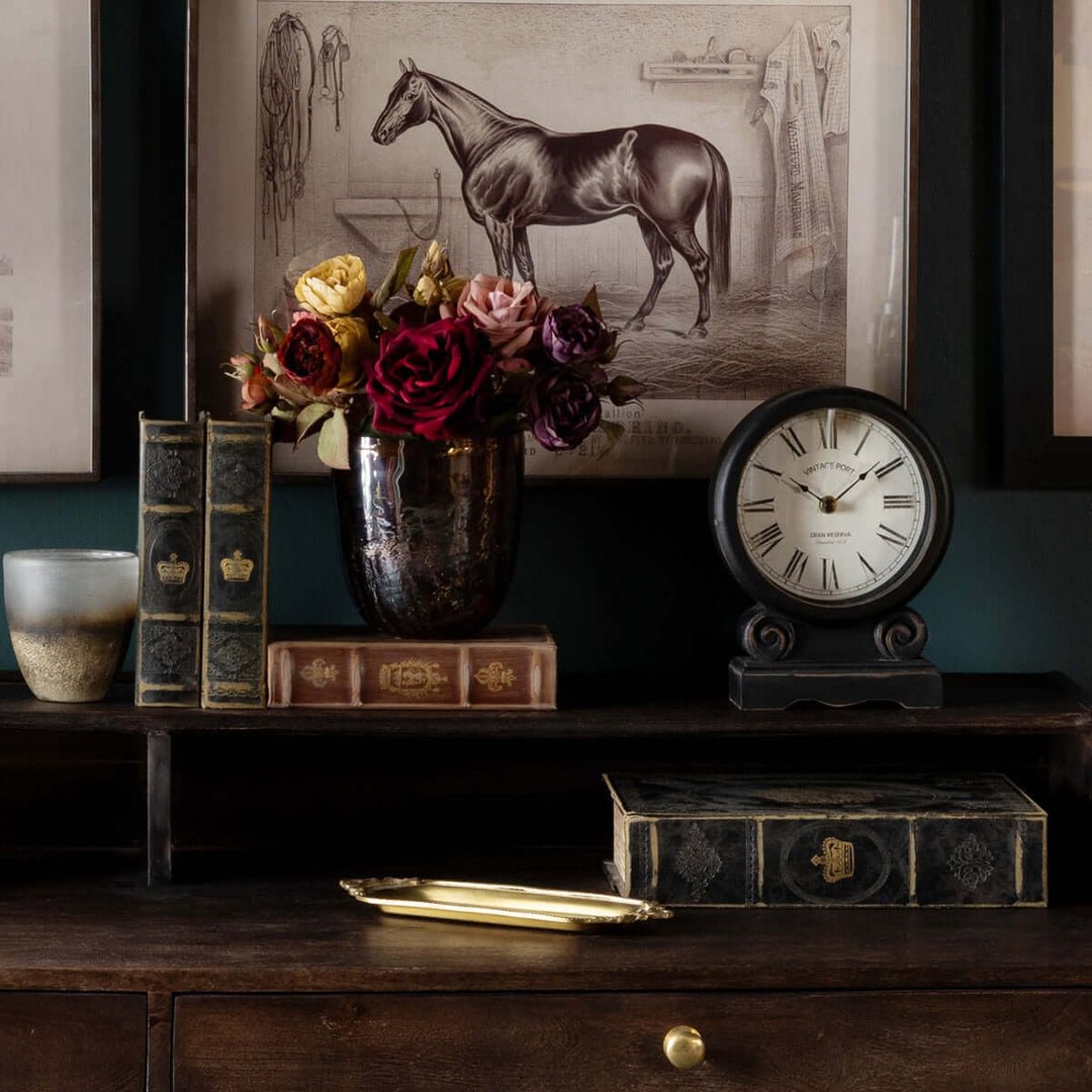 Antique-style Roman numeral mantel clock in black finish on a vintage shelf with books and flowers.