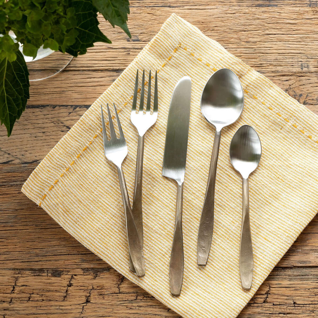 Hand-forged antique gold flatware set on a yellow napkin, including forks, knife, and spoons, displayed on a rustic wooden table.