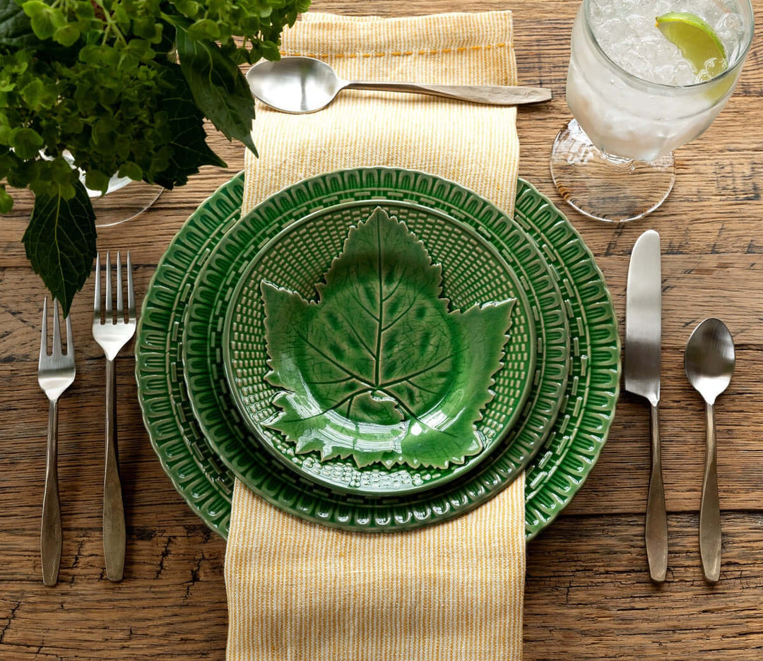 Table setting with green leaf-patterned plates and antique flatware on a rustic wooden table, accented by a yellow napkin.