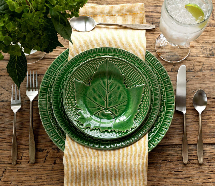 Table setting with green leaf-patterned plates and antique flatware on a rustic wooden table, accented by a yellow napkin.