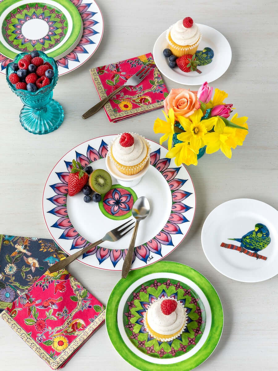 Vibrant table setting with floral plates, cupcakes, antique flatware, colorful napkins, and a vase of fresh flowers on a white table.
