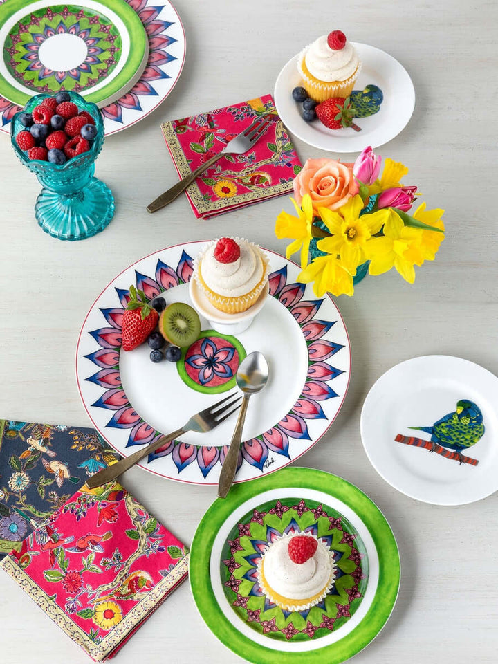 Vibrant table setting with floral plates, cupcakes, antique flatware, colorful napkins, and a vase of fresh flowers on a white table.