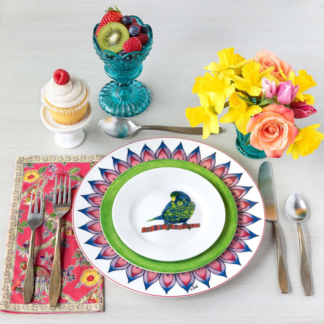 Colorful dining setup with a parrot-themed plate, antique gold flatware, vibrant flowers, fruit bowl, and cupcake.