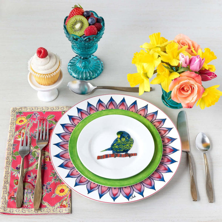 Colorful dining setup with a parrot-themed plate, antique gold flatware, vibrant flowers, fruit bowl, and cupcake.