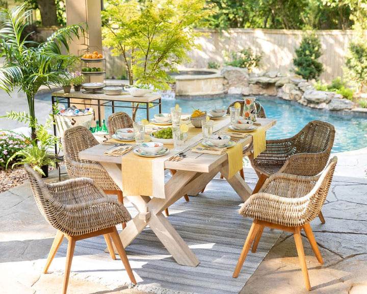 Elegant outdoor dining setup with handwoven chairs and a wooden table by a poolside, styled with vintage-inspired cutlery and decor.