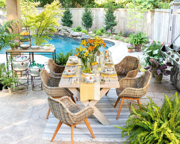 Outdoor dining setup with wicker chairs and a wooden table by a pool, adorned with vibrant flowers and elegant tableware.