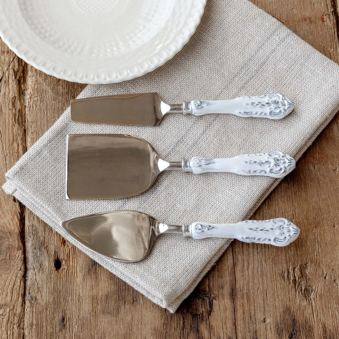 Embossed White Enamel Cheese Knife Set on rustic table, stainless steel blades with vintage-inspired aluminum handles on linen cloth.