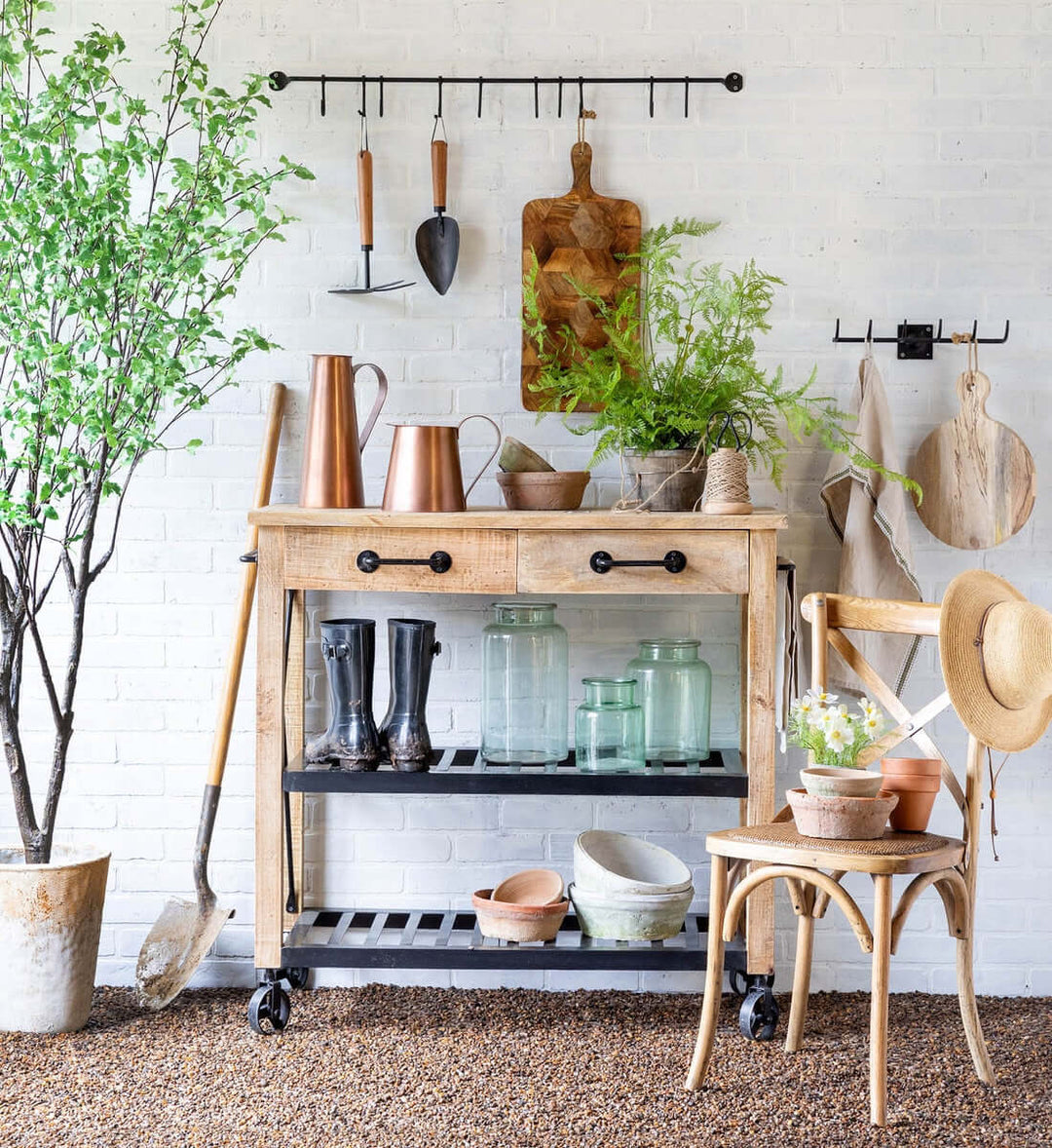 Vintage rolling garden cart with drawers, mango wood and black iron, styled with plants and tools against a white brick wall.