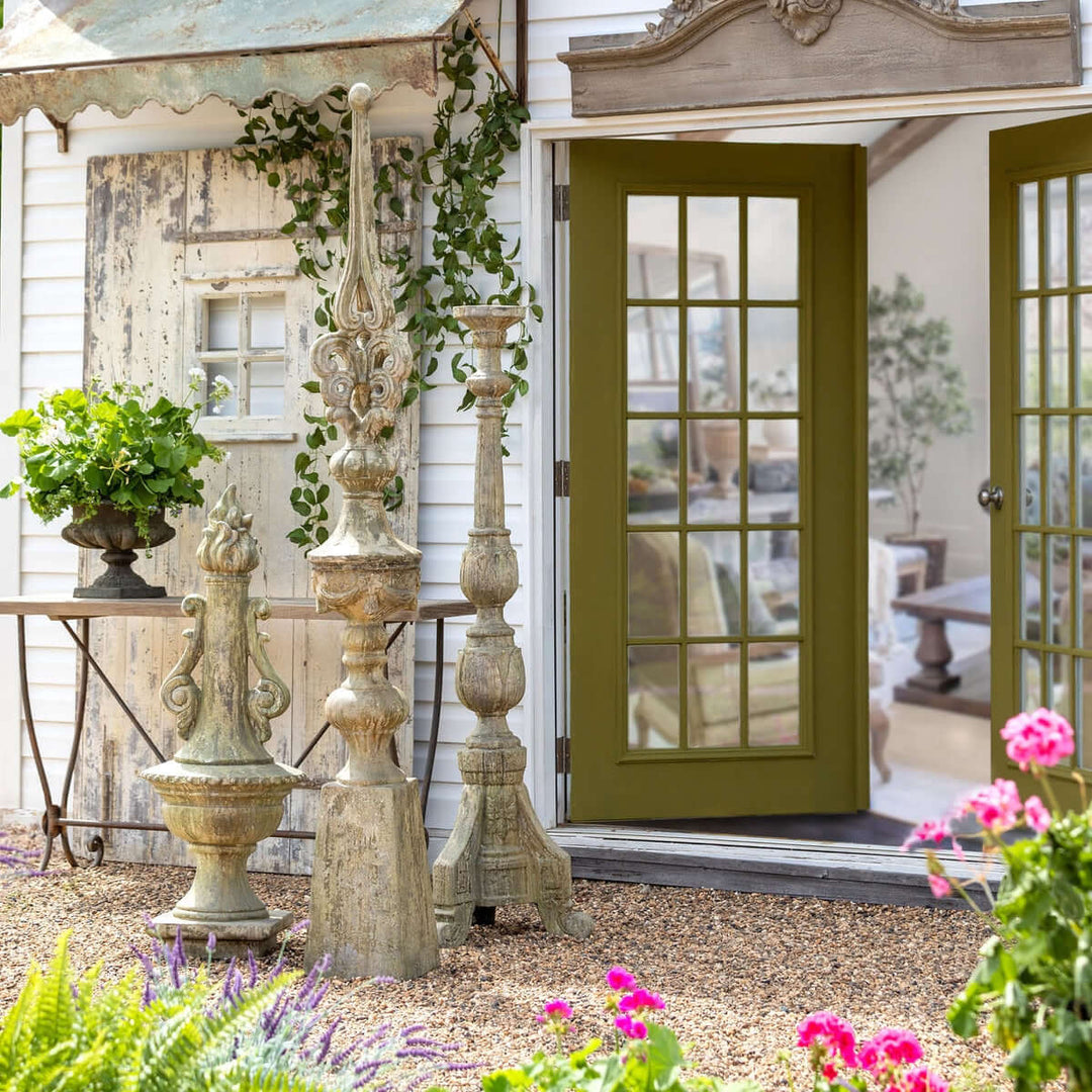 Antique Grecian garden torch statues with a rustic finish displayed near a cottage entrance adorned with plants and flowers.