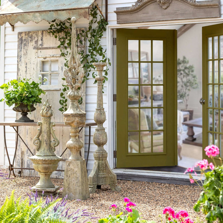 Antique Grecian garden torch statues with a rustic finish displayed near a cottage entrance adorned with plants and flowers.