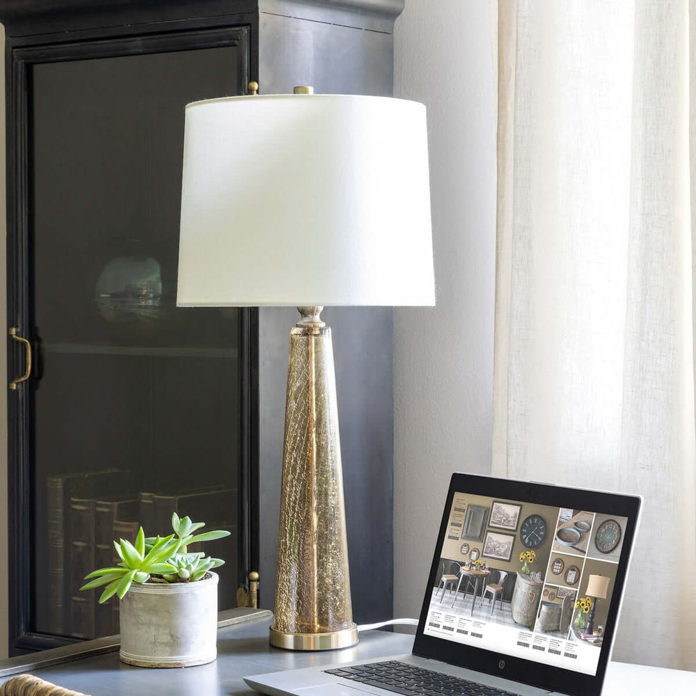 Crackled glass table lamp with white linen shade on desk beside laptop and potted plant, adding elegance and warmth to room decor.
