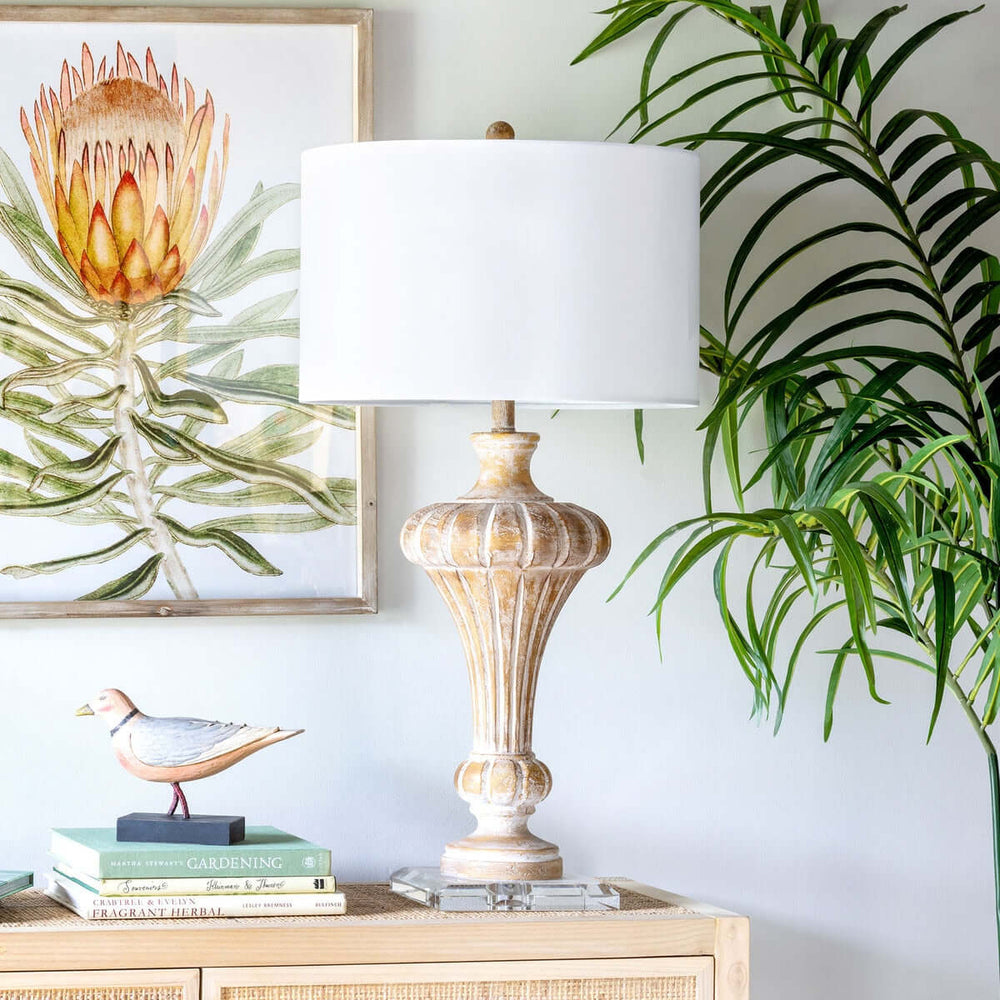 Vintage whitewashed urn table lamp with crystal base and linen shade on wooden table, decorative plant and art in the background.