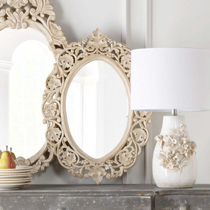 Vintage hand-carved wood wall mirror with intricate floral scrollwork and oval shape, next to a decorative vase on a rustic table.