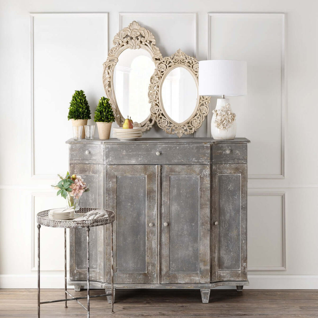 Vintage hand carved wood wall mirror with ornate floral design on a rustic cabinet, surrounded by potted plants and decor.