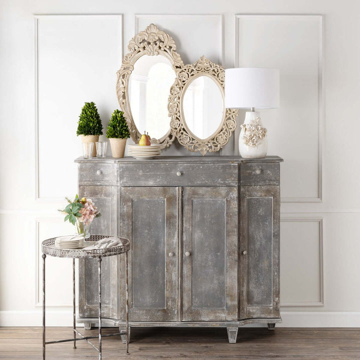Vintage hand carved wood wall mirror with ornate floral design on a rustic cabinet, surrounded by potted plants and decor.