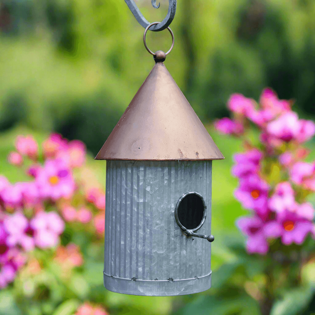 Rustic galvanized birdhouse with copper roof, hanging in a garden surrounded by colorful flowers.