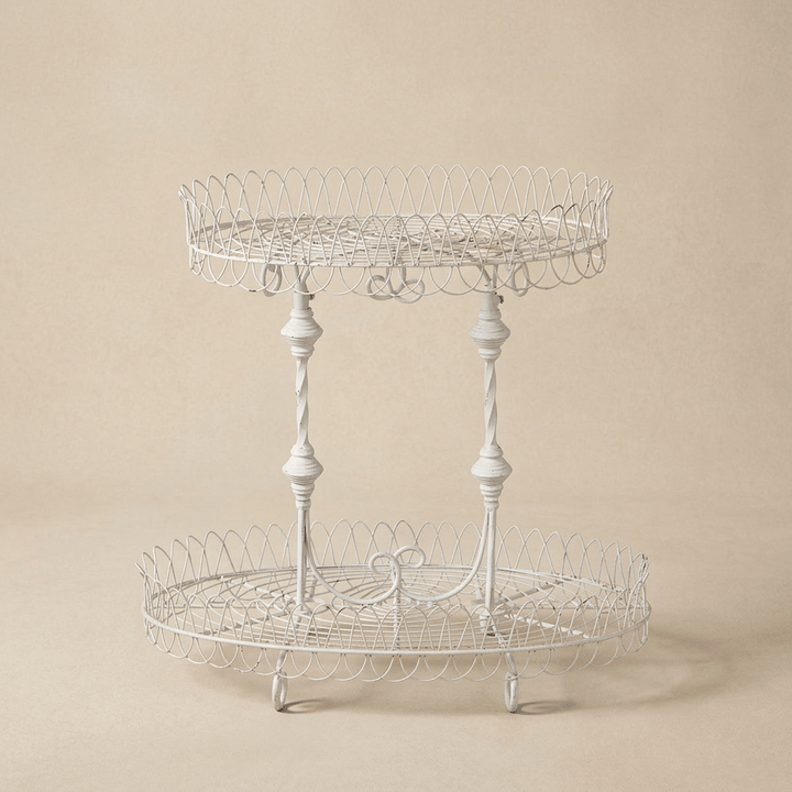 Fleury antiqued white iron plant stand with two oval wire shelves and decorative spindle supports against a warm beige background.
