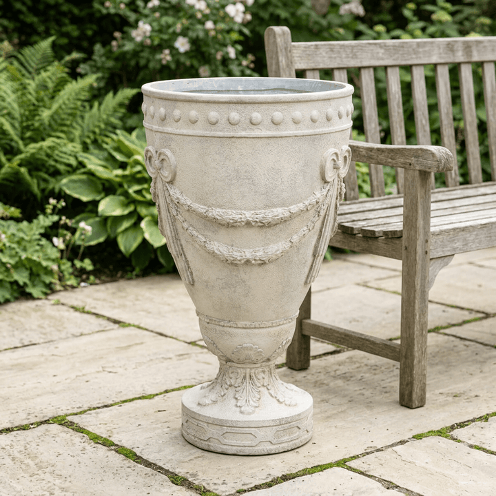 Florentine Floral Urn Planter in a weathered ivory finish on a stone patio beside a wooden garden bench.