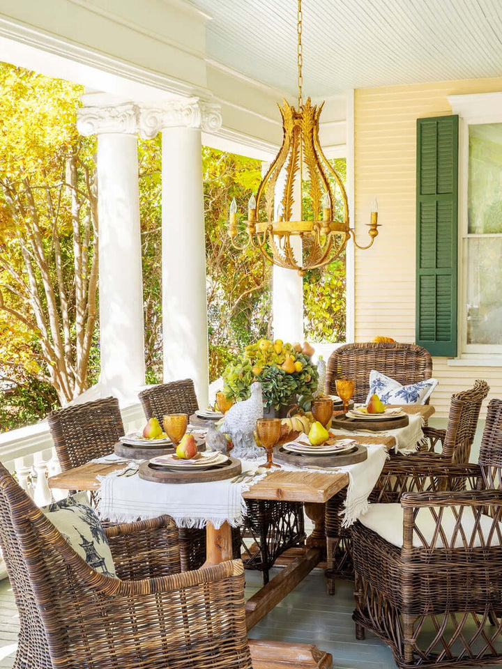 Outdoor dining area with wicker chairs, a wooden table, and a gold chandelier on a porch.