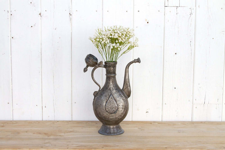 Decorative metal pitcher with flowers on a wooden surface and white wooden panel background
