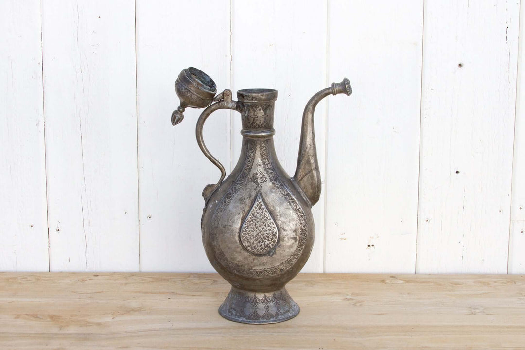 Decorative metal pitcher on a wooden surface with a white wooden panel background