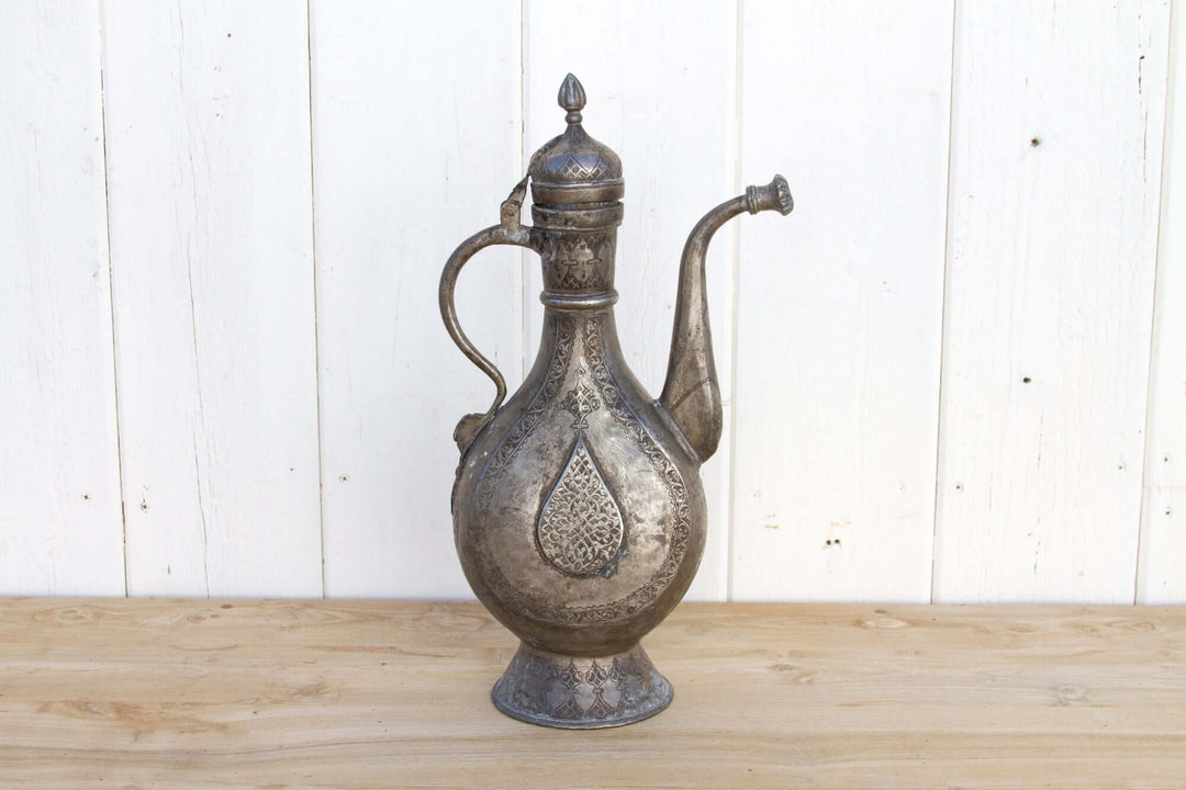 Decorative metal pitcher on a wooden surface with a white wooden panel background