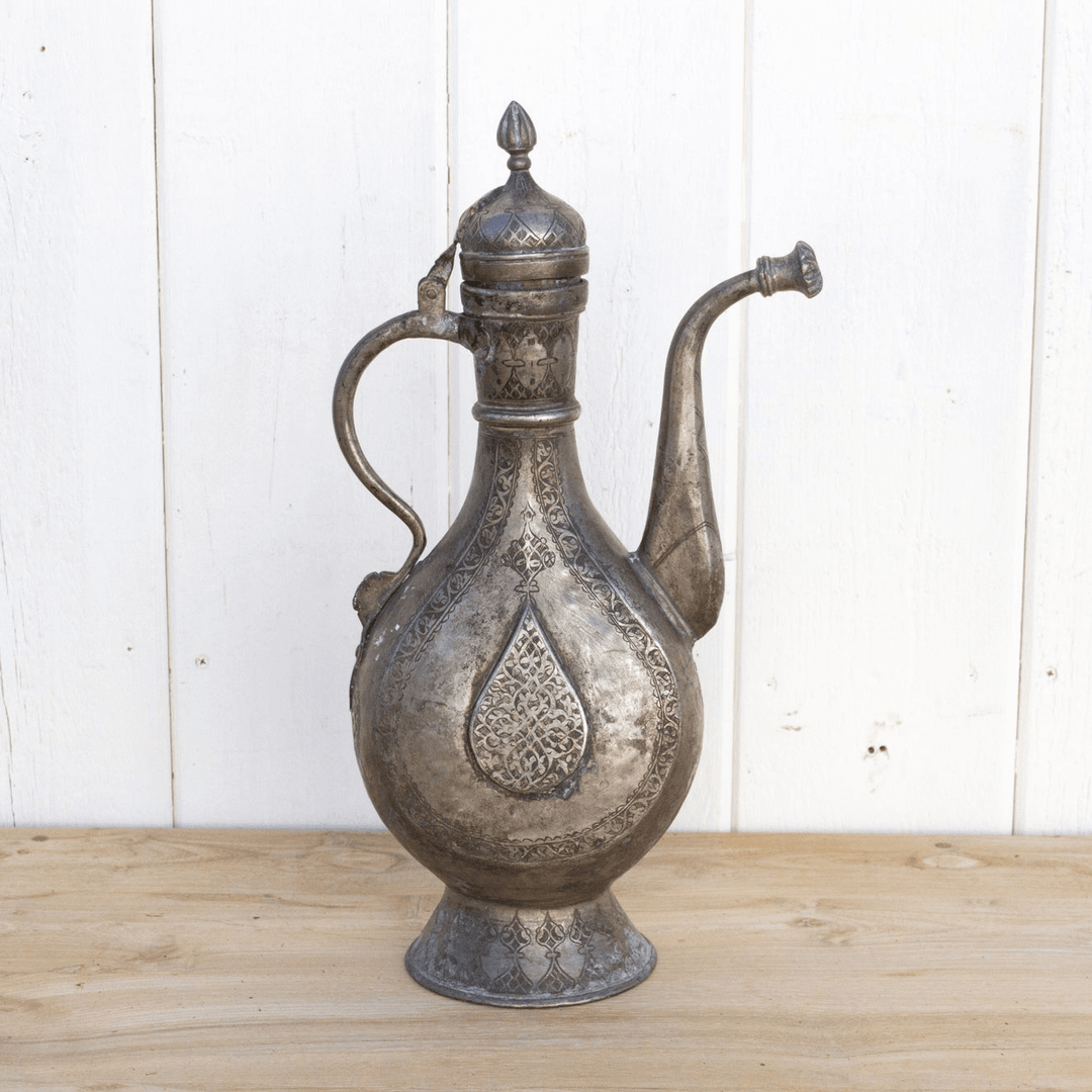 Decorative metal pitcher with intricate designs on a wooden surface and white wall background